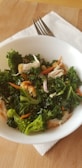 A white bowl filled with a fresh salad containing green kale, broccoli, shredded carrots, and pieces of chicken on a wooden surface. A fork rests on a white napkin beside the bowl.
