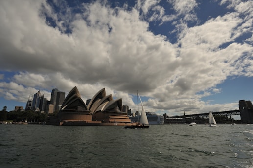 A large opera house with distinctive sail-like architecture is visible near the water's edge. Several sailboats and a few larger vessels are seen on the water in front. A bridge spans across the right side of the background, and modern skyscrapers are observed to the left, under a sky filled with large, fluffy clouds.