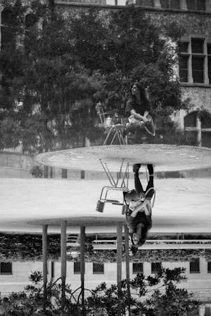 A black and white artistic photograph featuring a reflection of a person sitting on a chair in a pool of water. The person, wearing casual clothing, appears thoughtful while surrounded by trees and an old building with large windows.