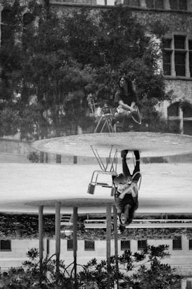 A black and white artistic photograph featuring a reflection of a person sitting on a chair in a pool of water. The person, wearing casual clothing, appears thoughtful while surrounded by trees and an old building with large windows.