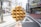 A close-up of a waffle on a stick with chocolate drizzle held by a hand in the foreground. The background features a street scene with a few barren trees and multiple buildings, including storefronts and urban architecture. The sky appears bright, suggesting daytime.