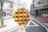 A close-up of a waffle on a stick with chocolate drizzle held by a hand in the foreground. The background features a street scene with a few barren trees and multiple buildings, including storefronts and urban architecture. The sky appears bright, suggesting daytime.