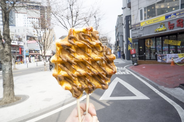 A close-up of a waffle on a stick with chocolate drizzle held by a hand in the foreground. The background features a street scene with a few barren trees and multiple buildings, including storefronts and urban architecture. The sky appears bright, suggesting daytime.