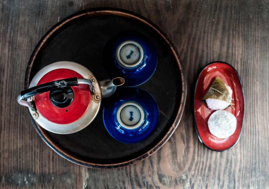 A metal teapot with a red lid is placed on a circular wooden tray along with two blue ceramic teacups. To the right side of the tray, there is a red oval plate with two pieces of traditional Japanese sweets, one wrapped in a leaf.