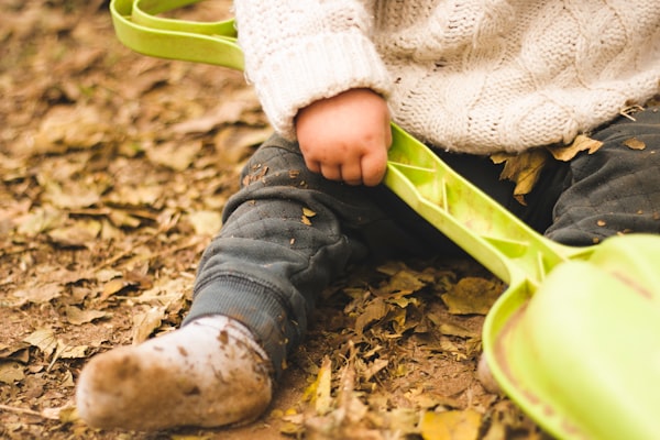 a young child wearing a cable-knit sweater and dirty socks sitting on leaf-covered dirt and holding a toy shovel