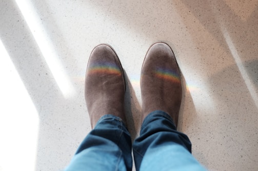 A pair of suede boots with a colorful rainbow reflection on them, standing on a light-colored speckled floor. The legs wearing the boots are clad in blue jeans.
