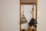 A minimalistic wall-mounted organizer holding two cleaning tools, a dustpan with a brush and a feather duster. The objects are hanging against a plain wall with a light switch nearby, framed by a simple wooden structure.