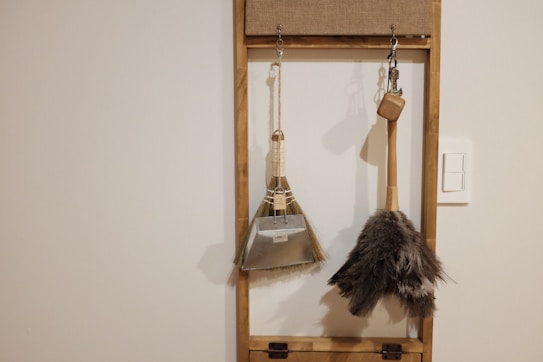 A minimalistic wall-mounted organizer holding two cleaning tools, a dustpan with a brush and a feather duster. The objects are hanging against a plain wall with a light switch nearby, framed by a simple wooden structure.