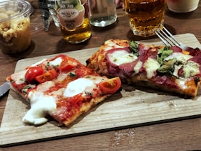 Two rectangular slices of pizza are presented on a wooden board. One slice is topped with fresh tomatoes, basil, and melted mozzarella cheese, while the other slice features cured meat, basil, and cheese. Nearby, there are drinks, including a glass of amber liquid and a bottle labeled apple juice, as well as a small container of crumble.