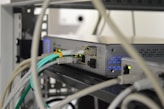 A close-up of a network switch or server rack with various colored Ethernet cables plugged into ports. The device features indicator lights, and the setting appears to be a technical or IT environment with several wires and cables.