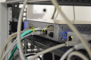 A close-up of a network switch or server rack with various colored Ethernet cables plugged into ports. The device features indicator lights, and the setting appears to be a technical or IT environment with several wires and cables.