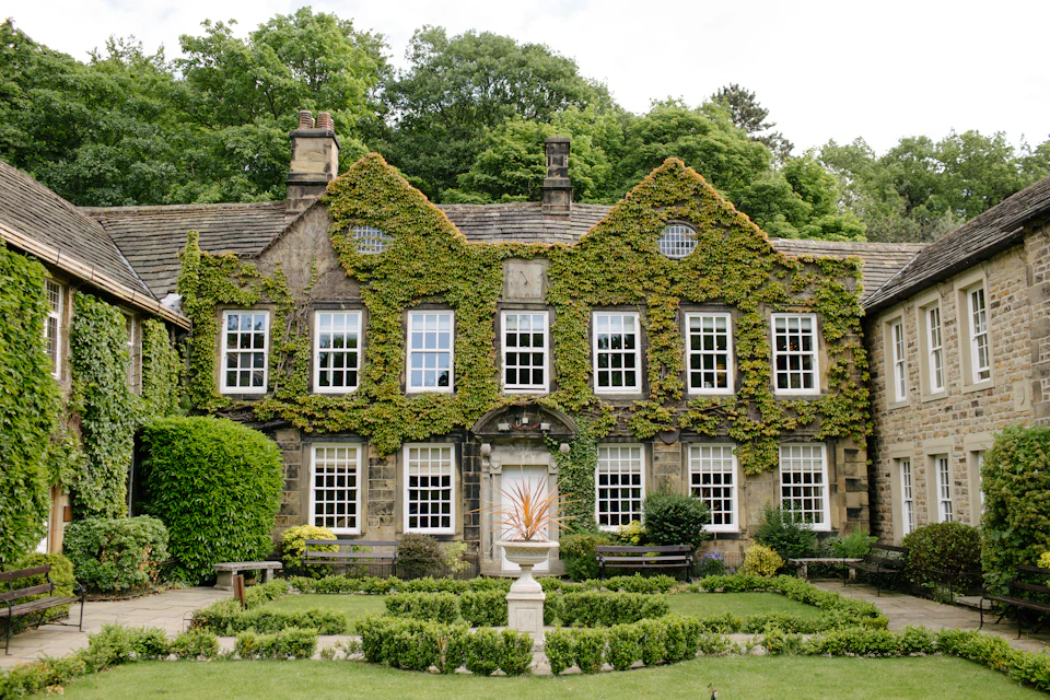 The image depicts a large, two-story stone house with a green roof and white walls nestled amidst lush greenery. The house features numerous windows that reflect the surrounding nature, and it is surrounded by an abundance of trees and bushes. In front of the house, there are several benches arranged in a circular pattern, inviting visitors to sit and enjoy the serene surroundings.