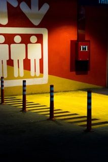 The image features a section of a parking garage or similar space with a vivid yellow floor and a large graphic on a red wall depicting three stylized human figures standing next to an arrow pointing downwards. Several red and blue traffic bollards stand in a row along the ground, casting distinct shadows due to bright overhead lighting. A small red box, possibly a fire alarm or similar equipment, is mounted on the wall.