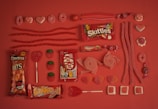 An assortment of red and pink candies are arranged neatly on a matching red background. The selection includes Skittles, Kit Kat, Doritos Bits, Trident gum, various gummies, licorice, lollipops, and chocolate bars. The candies are organized in rows, displaying different shapes and textures, creating a visually appealing pattern.