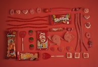 An assortment of red and pink candies are arranged neatly on a matching red background. The selection includes Skittles, Kit Kat, Doritos Bits, Trident gum, various gummies, licorice, lollipops, and chocolate bars. The candies are organized in rows, displaying different shapes and textures, creating a visually appealing pattern.