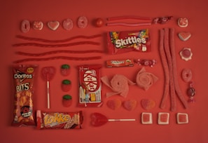 An assortment of red and pink candies are arranged neatly on a matching red background. The selection includes Skittles, Kit Kat, Doritos Bits, Trident gum, various gummies, licorice, lollipops, and chocolate bars. The candies are organized in rows, displaying different shapes and textures, creating a visually appealing pattern.