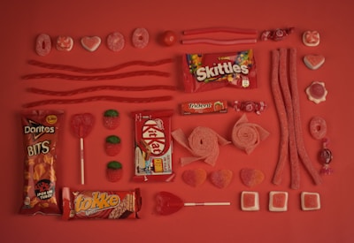 An assortment of red and pink candies are arranged neatly on a matching red background. The selection includes Skittles, Kit Kat, Doritos Bits, Trident gum, various gummies, licorice, lollipops, and chocolate bars. The candies are organized in rows, displaying different shapes and textures, creating a visually appealing pattern.