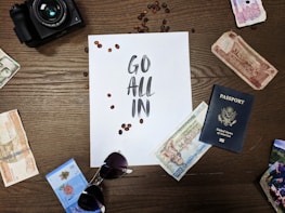 A wooden table displays various travel-related items, including international currency notes, a U.S. passport, a pair of sunglasses, and a camera. In the center is a sheet of paper with the words 'Go All In' surrounded by scattered coffee beans.