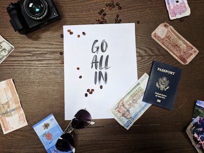 A wooden table displays various travel-related items, including international currency notes, a U.S. passport, a pair of sunglasses, and a camera. In the center is a sheet of paper with the words 'Go All In' surrounded by scattered coffee beans.