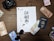 A wooden table displays various travel-related items, including international currency notes, a U.S. passport, a pair of sunglasses, and a camera. In the center is a sheet of paper with the words 'Go All In' surrounded by scattered coffee beans.