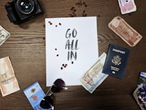 A wooden table displays various travel-related items, including international currency notes, a U.S. passport, a pair of sunglasses, and a camera. In the center is a sheet of paper with the words 'Go All In' surrounded by scattered coffee beans.
