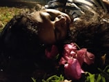A person lies on the ground with their eyes closed, surrounded by pink flowers and grass. The lighting is dim, creating a peaceful and serene atmosphere.
