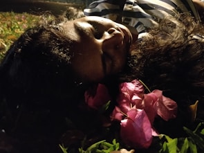 A person lies on the ground with their eyes closed, surrounded by pink flowers and grass. The lighting is dim, creating a peaceful and serene atmosphere.