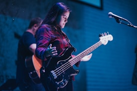 A musician is playing an electric bass guitar while standing on stage. The person is wearing a red plaid shirt and appears focused on the music. A microphone is visible to the right, and another person with a guitar is in the background.