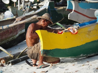 An older man wearing a straw hat is sitting on the sandy beach while repairing or working on a colorful wooden boat. The boat is painted with vibrant yellow and green colors, and there are other boats and fishing equipment in the background.