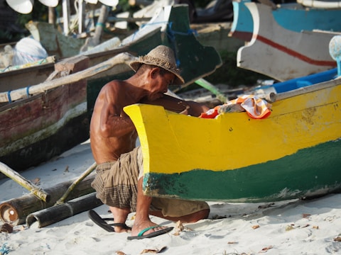 An older man wearing a straw hat is sitting on the sandy beach while repairing or working on a colorful wooden boat. The boat is painted with vibrant yellow and green colors, and there are other boats and fishing equipment in the background.