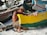 An older man wearing a straw hat is sitting on the sandy beach while repairing or working on a colorful wooden boat. The boat is painted with vibrant yellow and green colors, and there are other boats and fishing equipment in the background.