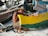 An older man wearing a straw hat is sitting on the sandy beach while repairing or working on a colorful wooden boat. The boat is painted with vibrant yellow and green colors, and there are other boats and fishing equipment in the background.