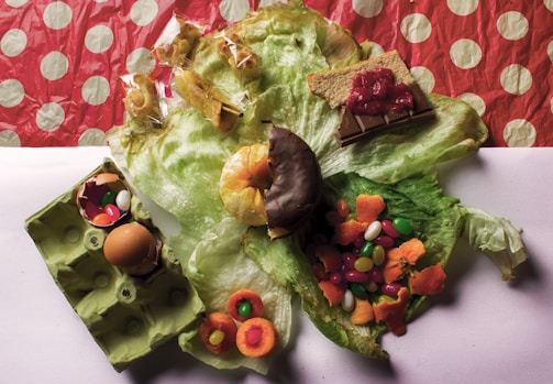 A variety of food items are arranged over a large piece of lettuce. There are jellybeans, an egg in an egg carton, orange peels, packaged muffins, chocolate bar with a graham cracker and jelly, a chocolate-covered donut, and candies. The background has a red polka-dot pattern.