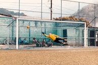 A soccer goalkeeper in a yellow jersey makes an impressive dive to attempt saving a ball that is heading towards the top corner of the goal. Several people sit behind the goal, watching the action unfold, with a bicycle positioned to the side. The scene is set against an outdoor soccer field with a net fence and mountainous background.