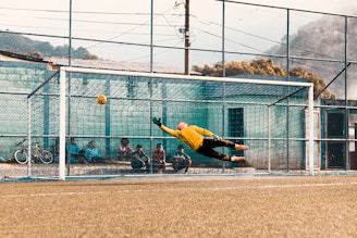A soccer goalkeeper in a yellow jersey makes an impressive dive to attempt saving a ball that is heading towards the top corner of the goal. Several people sit behind the goal, watching the action unfold, with a bicycle positioned to the side. The scene is set against an outdoor soccer field with a net fence and mountainous background.