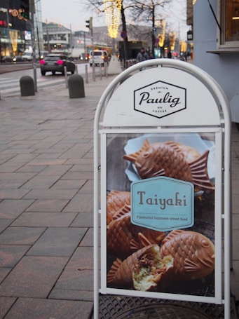 A street advertisement signboard displays the brand 'Paulig' and features an image of Taiyaki, which is a fish-shaped Japanese street food pastry, positioned on a city sidewalk with a backdrop of urban traffic and illuminated trees.