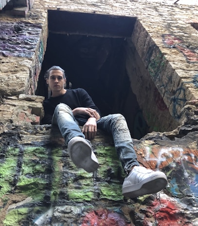 A person is sitting casually on a weathered, graffiti-covered stone wall, wearing a black shirt and distressed jeans. Their white sneakers stand out as they dangle their legs over the edge. The setting appears to be an old, possibly abandoned building with a rough, gritty appearance.