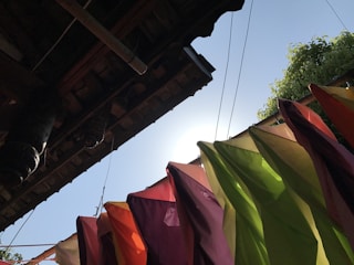 Colorful fabric pieces are hanging and fluttering in the breeze beneath the edge of a wooden roof. The sun is visible in the background, casting a glow through the vibrant cloth in shades of red, orange, purple, yellow, and green. A clear blue sky and some greenery are also present.