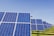 A field of solar panels arranged in rows under a clear blue sky, capturing sunlight to convert into energy. The panels are mounted on metal structures above a grassy area.