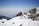 A person with a backpack is trekking on a snow-covered mountain ridge. The landscape features rugged mountain peaks and a clear blue sky in the background. The scene is serene with snow patches on the rocks and distant mountain ranges visible through a light mist.