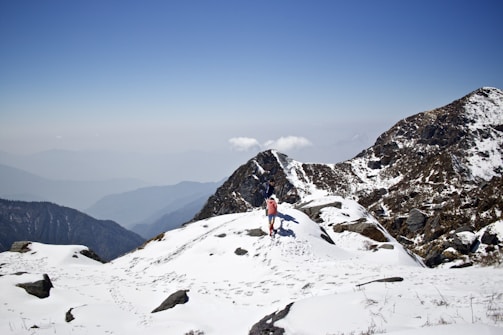 A person with a backpack is trekking on a snow-covered mountain ridge. The landscape features rugged mountain peaks and a clear blue sky in the background. The scene is serene with snow patches on the rocks and distant mountain ranges visible through a light mist.