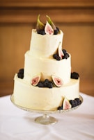 A three-tiered cake with a smooth cream-colored frosting is elegantly decorated with fresh figs and blackberries arranged artistically on each layer. The cake is presented on a delicate, reflective cake stand, set against a soft background.