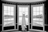A white wedding dress hangs elegantly on a hanger in front of three large, arched windows. The windows overlook a serene outdoor scene with lush greenery and a river. The setting is indoors, likely in a formal room or venue.