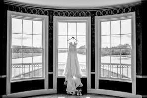 A white wedding dress hangs elegantly on a hanger in front of three large, arched windows. The windows overlook a serene outdoor scene with lush greenery and a river. The setting is indoors, likely in a formal room or venue.