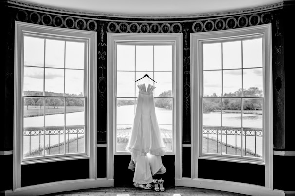 A white wedding dress hangs elegantly on a hanger in front of three large, arched windows. The windows overlook a serene outdoor scene with lush greenery and a river. The setting is indoors, likely in a formal room or venue.