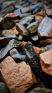A collection of wet stones, varying in size and shape, predominantly in shades of brown, gray, and black. The surface of the stones appears smooth, likely due to water exposure, giving them a glistening appearance.