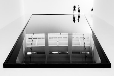 A large, reflective black surface lies in a minimalistic gallery space, capturing the reflection of two standing figures and the ceiling above. The setting is stark with high contrast, dominated by clean lines and an austere aesthetic.