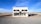 A small standalone building resembling a Prada storefront is situated in an isolated desert landscape. The structure has a minimalist design, featuring large display windows containing a few luxury bags and shoes. The sky is vast and clear, complementing the remote setting, and a dirt road stretches in front of the building.