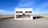 A small standalone building resembling a Prada storefront is situated in an isolated desert landscape. The structure has a minimalist design, featuring large display windows containing a few luxury bags and shoes. The sky is vast and clear, complementing the remote setting, and a dirt road stretches in front of the building.