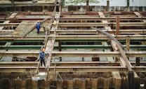 Workers in protective gear are seen on a construction site characterized by metal beams, scaffolding, and wooden supports. They appear to be engaged in construction tasks on a large infrastructure project, possibly a bridge or a similar structure.